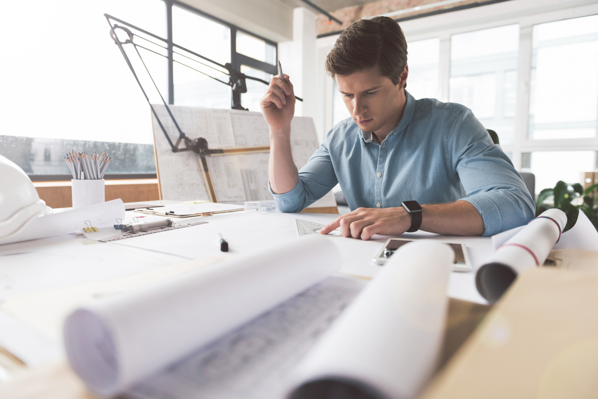 Professional architect is working in office Involved in project. Serious qualified young engineer is drawing blueprint. He is sitting at desk and holding pencil while looking at paper thoughtfully. Copy space in the left side